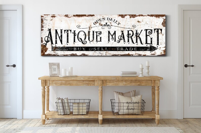 May include: A rustic white sign with black lettering that reads "Open Daily Antique Market Buy Sell Trade" with a black arrow pointing to the right. The sign is hanging above a wooden console table with two black wire baskets and a throw pillow with the text "Welcome Home".