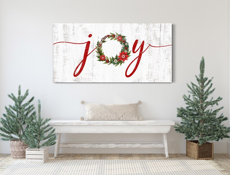 May include: White wooden sign with the word "joy" written in red cursive lettering. A red and green Christmas wreath with red berries and white flowers is incorporated into the letter "o".