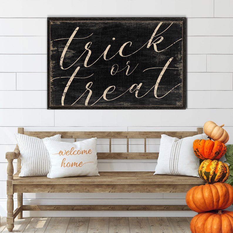 K&ouml;nnte beinhalten: Ein schwarzes Holzschild mit wei&szlig;en Buchstaben, auf dem "trick or treat" steht, h&auml;ngt an einer wei&szlig;en Schiffslappenwand. Eine Holzbank mit zwei Kissen und einem Stapel K&uuml;rbisse steht vor dem Schild.