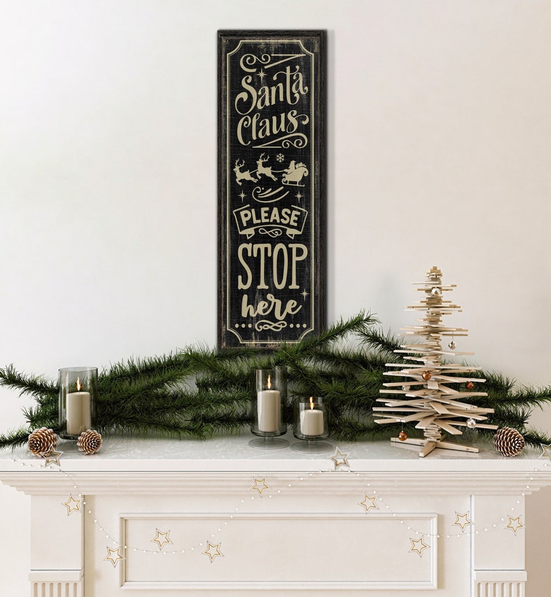 May include: A black wooden sign with white text that reads "Santa Claus Please Stop Here". The sign is decorated with a small image of Santa Claus in his sleigh with reindeer. The sign is hanging on a white wall above a fireplace mantel decorated with a small wooden Christmas tree, evergreen branches, and candles.