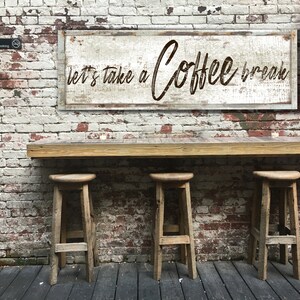 May include: A rustic wooden sign with white paint and brown lettering that reads "Let's take a Coffee break". The sign is hanging on a brick wall with a wooden counter and three wooden stools in front of it.
