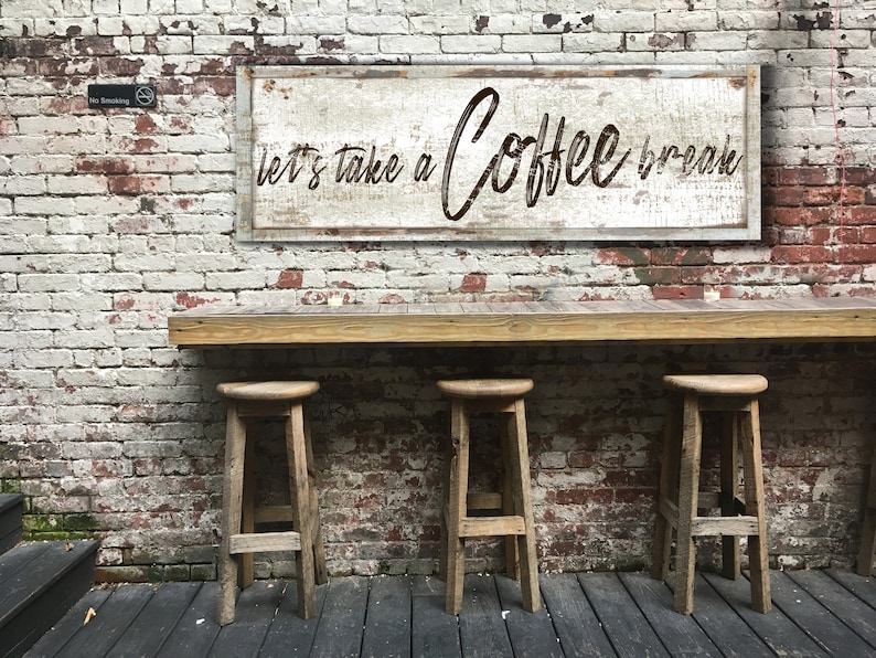 May include: A rustic wooden sign with white paint and brown lettering that reads "Let's take a Coffee break". The sign is hanging on a brick wall with a wooden counter and three wooden stools in front of it.