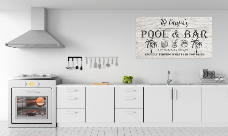 May include: A white wood sign with black text that reads "The Carson's Pool & Bar Est. 2017/Sarasota, FL Proudly Serving Whatever You Bring". The sign has palm trees and drink icons.