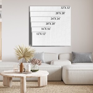 May include: A white canvas with different size options listed in inches. The canvas is hanging on a white wall in a living room setting with a white couch and a wooden coffee table.
