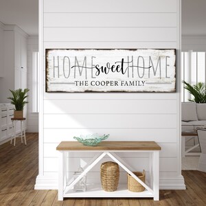 May include: White wooden sign with black text that reads "Home Sweet Home" and "The Cooper Family". The sign is hanging on a white wall in a home interior setting.