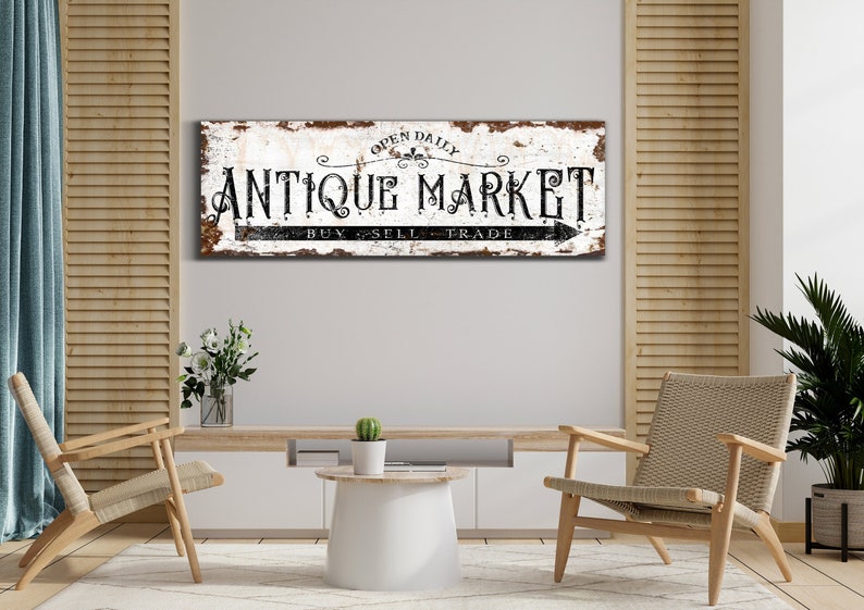 May include: A rustic white sign with black lettering that reads "Open Daily Antique Market Buy Sell Trade" with a black arrow pointing to the right. The sign is hanging on a white wall in a room with two chairs and a table.