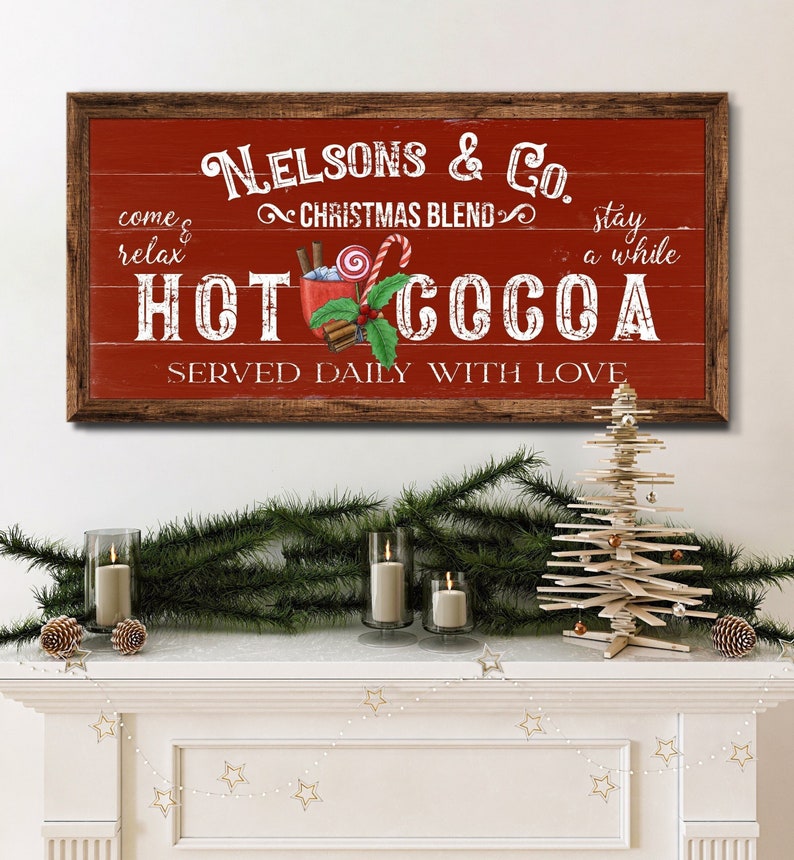 Op de afbeelding: Een rood houten bord met witte tekst die "Nelsons & Co. Christmas Blend Hot Cocoa Served Daily With Love" luidt. Het bord heeft een rustieke uitstraling en toont een cartoonafbeelding van een mok warme chocolademelk met marshmallows, snoepstokken en kaneelstokjes.