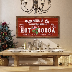 Op de afbeelding: Een rood en wit bord met de tekst "Nelsons & Co. Christmas Blend Hot Cocoa Served Daily With Love" en een afbeelding van een mok met warme chocolademelk met marshmallows, pepermuntstokken en kaneelstokjes.