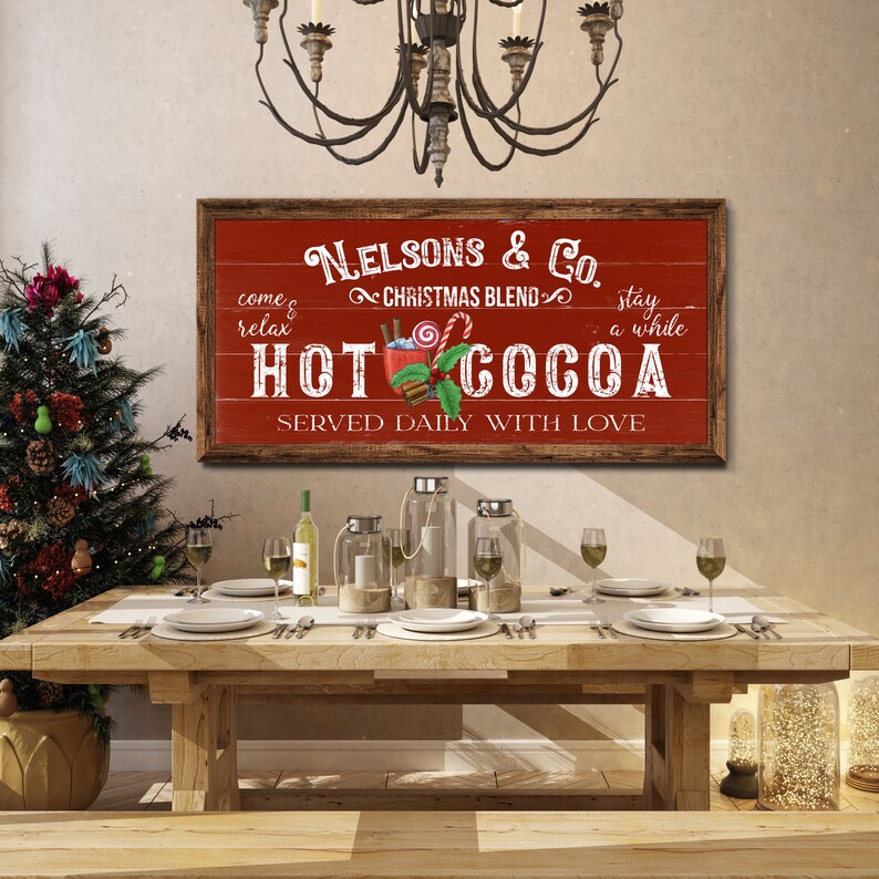 Op de afbeelding: Een rood en wit bord met de tekst "Nelsons & Co. Christmas Blend Hot Cocoa Served Daily With Love" en een afbeelding van een mok met warme chocolademelk met marshmallows, pepermuntstokken en kaneelstokjes.