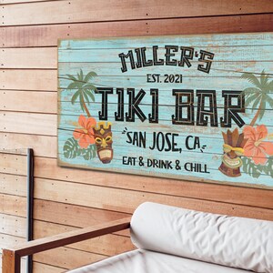 K&ouml;nnte beinhalten: Ein blaues und wei&szlig;es Holzschild mit dem Text "MILLER'S EST. 2021 TIKI BAR SAN JOSE, CA EAT & DRINK & CHILL". Das Schild zeigt eine Tiki-Maske, Palmen und tropische Blumen.
