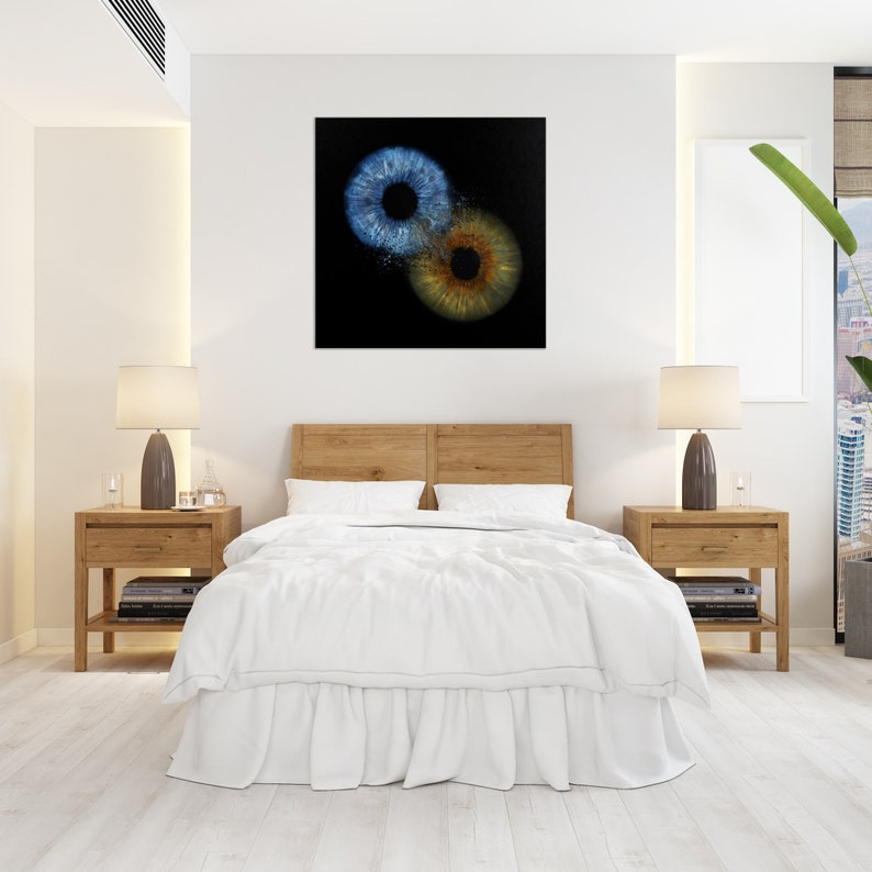 May include: A bedroom interior with a white bed and two nightstands. A framed artwork with two abstract eye-like shapes in blue and brown is hanging on the wall above the bed.