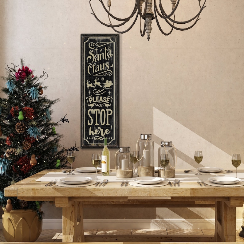 May include: A black wooden sign with white text that reads "Santa Claus Please Stop Here". The sign is hanging on a wall in a dining room with a wooden table set for a holiday dinner. There is a Christmas tree in the corner of the room.