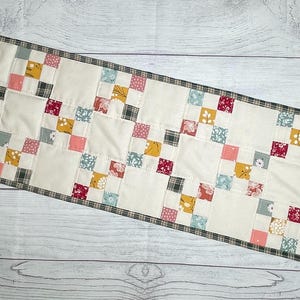 May include: A rectangular quilted table runner with a cream-colored background and a patchwork design. The patchwork features squares of various floral and plaid patterns in pink, yellow, red, and blue. The border is a plaid pattern.