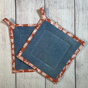 May include: Two denim square potholders with a rust-colored floral border and a loop for hanging. The potholders are made of blue denim fabric and are designed for kitchen use.
