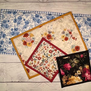 May include: A collection of floral-patterned fabric items, including placemats and pot holders. The placemats feature blue rose patterns and a gingham border. The pot holders showcase various floral designs with red, yellow, and pink flowers.