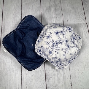May include: Two fabric bowl cozies. One is a dark blue denim, the other is white with a blue floral pattern. The cozies are square with rounded corners and are designed to hold bowls, protecting hands from heat.