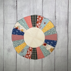 May include: A circular patchwork quilt with a light wood center. The quilt features a variety of fabrics in shades of blue, pink, yellow, and green, with floral and plaid patterns. The quilt is approximately 12 inches in diameter.