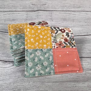 May include: A stack of square fabric coasters with a patchwork design. Each coaster features four squares of different floral patterns in yellow, white, pink, and teal. The coasters are made of fabric and have a stitched edge.