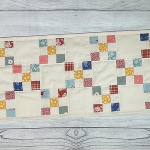May include: A rectangular quilted table runner with a cream-colored background. The runner features a repeating pattern of colorful squares in shades of red, blue, yellow, and pink, creating a patchwork design. The edges are neatly finished.