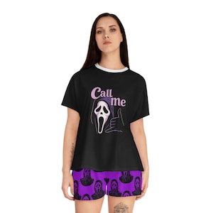 May include: A black t-shirt with a white crew neck and a graphic print featuring a purple hooded ghost mask making a hand gesture with the text Call me above it, paired with purple shorts featuring a repeating black ghost mask pattern.