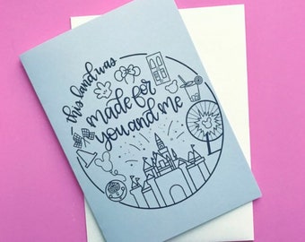 May include: A light blue card with a black outline of a castle and other Disney icons. The text on the card reads "This land was made for you and me."