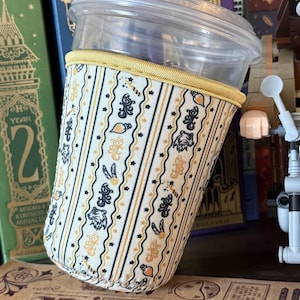 May include: A clear plastic cup with a fabric sleeve featuring a vertical striped pattern in cream, black, and gold. The fabric displays repeating icons of wolf heads, wands, and lightbulbs, finished with a gold-colored fabric trim at the top edge.