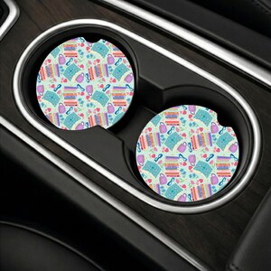 May include: Two circular car cup holder coasters featuring a light blue background with a repeating pattern of colorful stacked books, purple mugs, blue eyeglasses, small pink hearts, and tiny green leaves. Each coaster has a small notch on the edge.