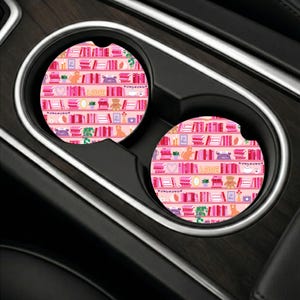 May include: Two circular car cup holder coasters featuring a repeating pattern of pink bookshelves filled with various books, small potted plants, a teddy bear, a typewriter, a camera, and the word LOVE printed in yellow on a light pink background.