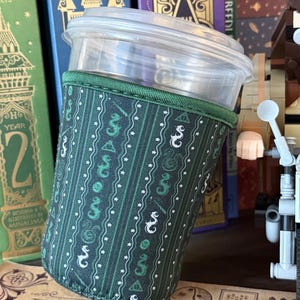 May include: A cylindrical cup sleeve featuring a dark green fabric with vertical stripes, small white stars, and repeating symbols including a triangle with a circle and line, and a serpent. The sleeve has a green fabric trim at the top edge.