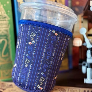 May include: A clear plastic cup with a dark blue fabric sleeve featuring a repeating pattern of gold snakes, small stars, and round glasses with red and yellow frames. The sleeve has a solid blue fabric trim along the top and bottom edges.