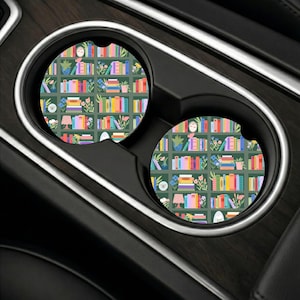 May include: Two circular car cup holder coasters featuring a repeating pattern of a dark green bookshelf filled with colorful books, potted plants, and small decorative items. Each coaster has a small notch on the edge for easy removal.