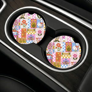 May include: Two circular car cup holder coasters featuring a colorful patchwork pattern. The design includes illustrations of white geese with red bows, various orange, pink, and yellow flowers, and checkered squares in shades of blue and pink.
