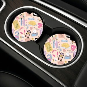 May include: Two round car cup holder coasters featuring a light pink background with a repeating pattern of books, a sleeping orange cat, pink headphones, a tissue box, a tea cup, and various literary-themed text labels and illustrations.