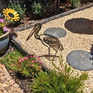 May include: Black metal garden art of a bird with a long beak standing on a bed of gravel. The bird is in silhouette and is surrounded by small stones.
