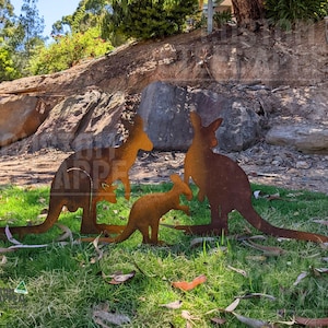 May include: A set of three rust-coloured metal kangaroo silhouettes standing on green grass. The group includes two larger adult kangaroos and one smaller joey kangaroo, all featuring a weathered, oxidised finish and flat, cutout profiles.