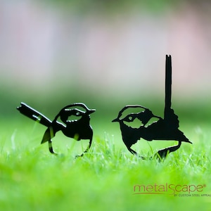 May include: Two black metal bird sculptures are set on green grass. The birds have detailed cut-out designs, with one bird's tail pointing upwards. The logo "metalscape" is visible at the bottom.