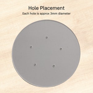 Puede incluir: Un objeto circular gris con cinco peque&ntilde;os agujeros espaciados uniformemente. El texto "Hole Placement" y "Each hole is approx 3mm diameter" son visibles. El objeto est&aacute; sobre una superficie de madera clara.