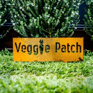 May include: A rusty yellow metal sign with the words "Veggie Patch" cut out. The sign is sitting on a bed of green grass.