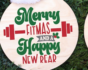 May include: Two round wooden signs with Christmas-themed messages. The first sign has a red Santa hat and says "Merry Fitmas and a Happy New Rear." The second sign has a gold tree and red bows and says "Merry Fitmas".