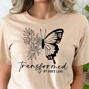 May include: A beige t-shirt with a black butterfly and floral design. The text "Transformed by God's Love" is printed on the shirt.