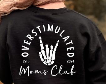 Op de afbeelding: Zwart sweatshirt met een witte afbeelding van een skelethand die het rock-and-roll-teken maakt in een cirkel met de tekst "Overstimulated Moms Club Est. 2024"