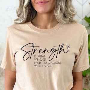 May include: A beige t-shirt with the text "Strength is what we gain from the madness we survive." in black lettering with a small heart symbol.