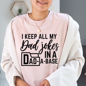 Op de afbeelding: Een roze T-shirt met de tekst "I keep all my Dad jokes in a Dad-a-base" in zwart gedrukt.
