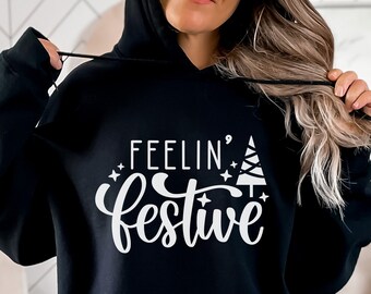 Puede incluir: Sudadera con capucha negra con texto blanco que dice "Feelin' Festive" con un gráfico de árbol de Navidad estilizado.