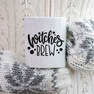 Op de afbeelding: Witte keramische mok met zwarte tekst die "Witches Brew" luidt met drie zwarte stippen aan weerszijden van de tekst.