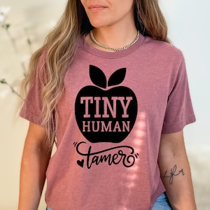 Puede incluir: Camiseta de color malva con un gráfico de manzana negra. El diseño de la manzana contiene las palabras "TINY HUMAN Tamer" en una fuente lúdica. La camiseta está hecha de un material suave.