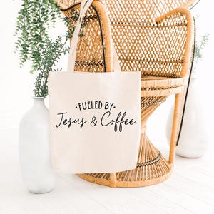 May include: A white canvas tote bag with black text that reads "Fueled by Jesus & Coffee". The bag is sitting on a wicker chair.
