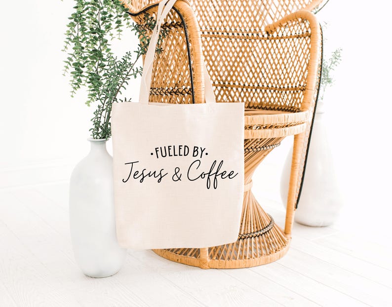 May include: A white canvas tote bag with black text that reads "Fueled by Jesus & Coffee". The bag is sitting on a wicker chair.
