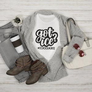 May include: White t-shirt with black text that reads "got ice? #schoolnurse". The shirt is laying on a gray cardigan with brown boots and a white bag.