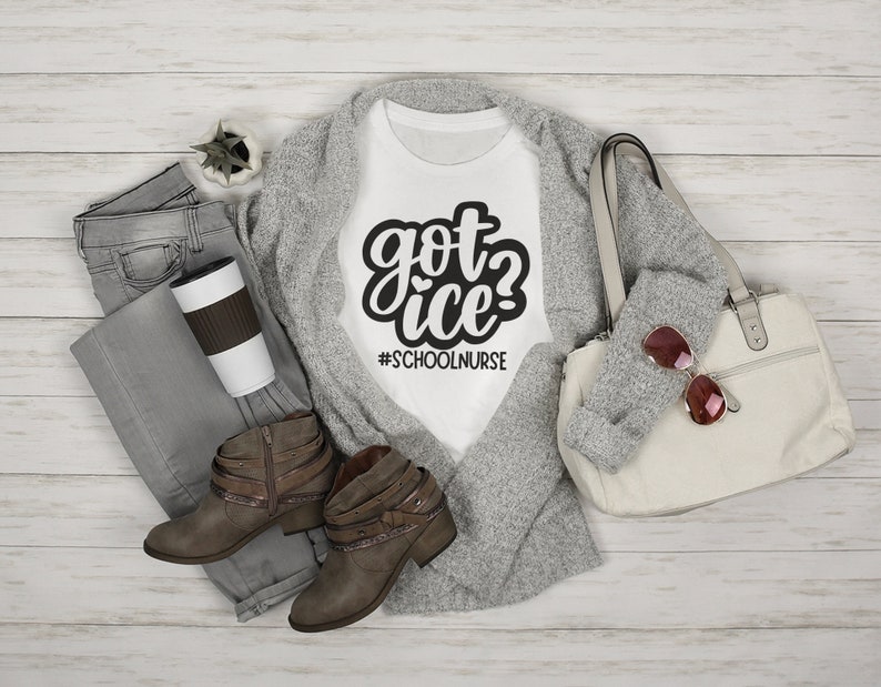 May include: White t-shirt with black text that reads "got ice? #schoolnurse". The shirt is laying on a gray cardigan with brown boots and a white bag.
